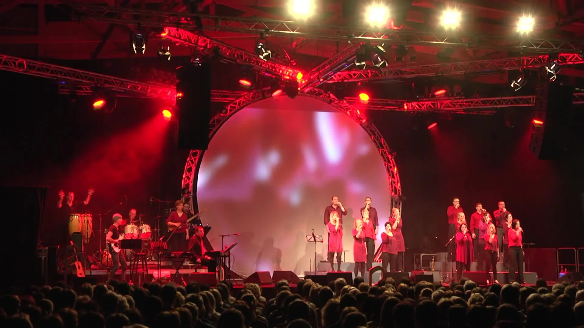 Konzert: Chor und Band auf der Bühne unter roten Scheinwerfern, davor das Publikum